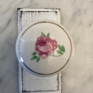 May include: White ceramic knob with a gold trim and a pink rose with green leaves design. The knob is attached to a white painted wooden base.