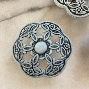 May include: A decorative metal knob with a floral design in a light blue colour. The knob has a circular centre and a scalloped edge.