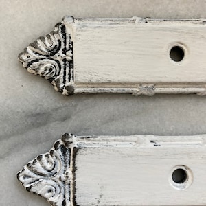 Peut inclure: Trois plaques en métal peintes en blanc avec des bords noirs vieillis et un trou au centre. Les plaques ont un motif décoratif dans le coin supérieur gauche.