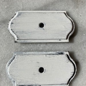 May include: Two white wooden plaques with a distressed finish and a round hole in the centre. The plaques have a slightly curved top and bottom edge.