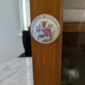 May include: A close-up of a cabinet door knob. The knob is round with a white background and a floral design featuring pink roses and gold accents. The knob is attached to a wooden cabinet door.