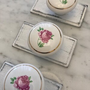 May include: Three white ceramic knobs with a gold trim and a pink rose design. The knobs are on a white painted wooden surface.