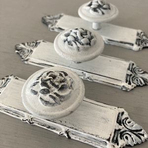 Peut inclure: Trois poignées de tiroir blanches avec des accents noirs. Chaque poignée présente un motif floral en relief et une base rectangulaire.