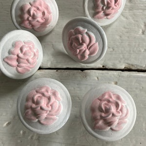 May include: Six white ceramic knobs with pink raised floral designs. The knobs are round and have a smooth surface.