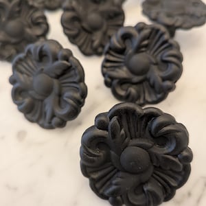 Peut inclure: Gros plan de plusieurs boutons de meubles floraux noirs mats. Les boutons présentent un design orné et détaillé avec un élément central circulaire et des feuilles stylisées. Les boutons sont disposés sur une surface en marbre blanc.
