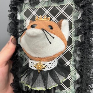 May include: A plush fox head with a gold crown and a star pendant, set against a white grid background. The fox wears a black and green ruffled collar. The frame is decorated with black lace.