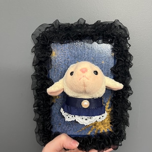 May include: A soft toy lamb in a navy blue dress with a pearl button, set against a starry blue backdrop. The artwork is framed with a black ruffled trim, creating a whimsical and decorative piece.