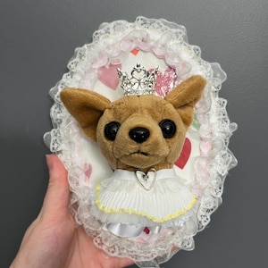May include: A plush Chihuahua wearing a silver crown and a white ruffled collar with a heart pendant. The dog is set against a heart-patterned background within a decorative oval frame with lace trim.