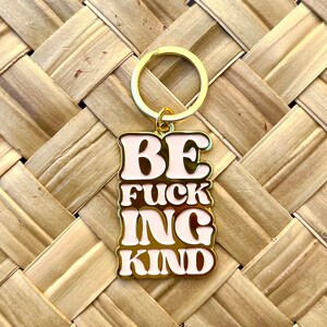 May include: Gold-colored keychain with a pink enamel charm that says "BE  FUCKING KIND" in a retro font.