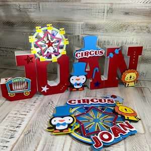 May include: Red, white, and blue wooden letters spelling "JOAN" with a circus theme. The letters are decorated with stars, a penguin, a lion, and a Ferris wheel. The Ferris wheel has a red star in the center and is surrounded by yellow and blue stripes.