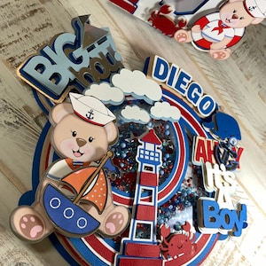 May include: A blue and red nautical themed cake topper with a teddy bear wearing a sailor hat, a lighthouse, a sailboat, and the words "Big Boy", "Diego", "Ahoy It's A Boy".