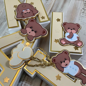 May include: Three white boxes shaped like the letters M, A, and I, decorated with gold trim and stars. Each box has a brown teddy bear with a blue shirt and a bow tie. The boxes are on a light wood surface.