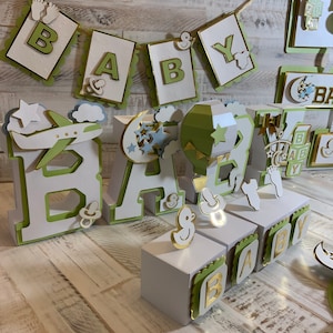 May include: Green and white 'BABY' banner with gold accents.  White wooden letters with green trim spelling 'BABY' with a green airplane, hot air balloon, and moon and stars decorations.  White boxes with green trim and gold letters spelling 'BABY'.