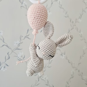 Könnte beinhalten: Ein gehäkeltes Hasenspielzeug, das einen rosa Ballon hält. Der Hase ist grau und hat schwarze Augen. Der Ballon ist mit einer Schnur an der Hand des Hasen befestigt.
