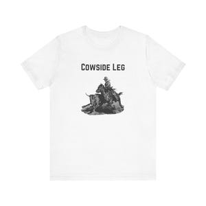 May include: White t-shirt featuring a black and white illustration of a cowboy on a horse roping a steer. The text "COWSIDE LEG" is printed above the graphic in a bold, sans-serif font. The shirt is made of a soft, lightweight material.