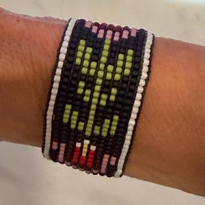 May include: A woven bracelet with a black background and a geometric design in green, pink, red, and white. The bracelet is worn on a wrist and features a repeating pattern of small beads.