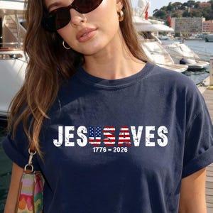 May include: A navy blue short-sleeve t-shirt featuring the white text JESUSA VES with the letters USA stylized as an American flag. Below the text, the numbers 1776-2026 are printed in white. The shirt has a crew neckline and a relaxed fit.