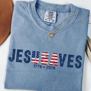 May include: A blue jean colored short-sleeve t-shirt with a crew neckline. The front features the dark blue text JESUSAVES with the letters USA filled with an American flag pattern. Below the text, the numbers 1776 - 2026 are printed in dark blue.