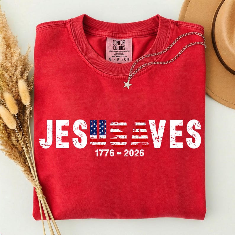 May include: A red crewneck t-shirt featuring the white distressed text "JESUS SAVES" with the letters "U" and "A" replaced by an American flag pattern. Below the text, the white numbers "1776 - 2026" are printed. The shirt has a Comfort Colors label.