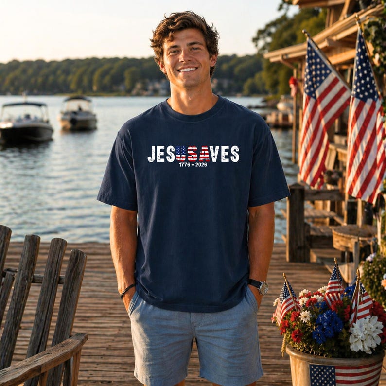 May include: A navy blue short-sleeve t-shirt featuring the white text JESUSA VES with the letters USA stylized as an American flag. Below the text, the numbers 1776-2026 are printed in white. The shirt is paired with light blue denim shorts.