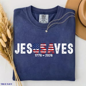 May include: A navy blue short-sleeve t-shirt featuring the white distressed text JESUS SAVES with the letters U and S stylized as an American flag. Below the text, the numbers 1776 - 2026 are printed in white. The shirt has a Comfort Colors brand label.