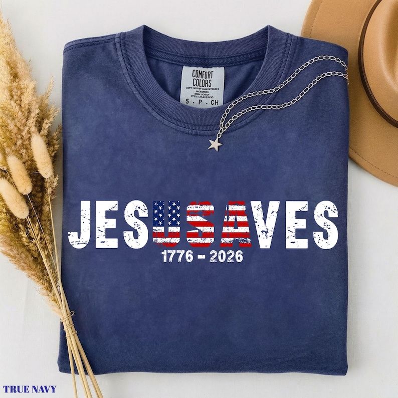 May include: A navy blue short-sleeve t-shirt featuring the white distressed text JESUS SAVES with the letters U and S stylized as an American flag. Below the text, the numbers 1776 - 2026 are printed in white. The shirt has a Comfort Colors brand label.