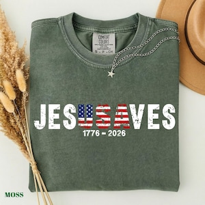 May include: A moss green short-sleeve t-shirt featuring the white distressed text JESUS SAVES with the letters U and S filled with an American flag pattern. Below the text, the numbers 1776 - 2026 are printed in white. A silver star necklace rests on the collar.