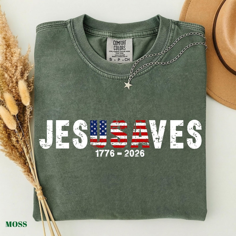 May include: A moss green short-sleeve t-shirt featuring the white distressed text JESUS SAVES with the letters U and S filled with an American flag pattern. Below the text, the numbers 1776 - 2026 are printed in white. A silver star necklace rests on the collar.