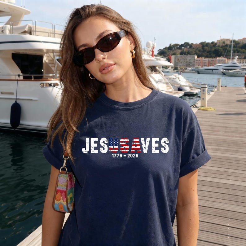 May include: A navy blue short-sleeve t-shirt featuring the white text JESUSA VES with the letters USA stylized as an American flag. Below the text, the numbers 1776-2026 are printed in white. The shirt has a crew neckline and a relaxed fit.