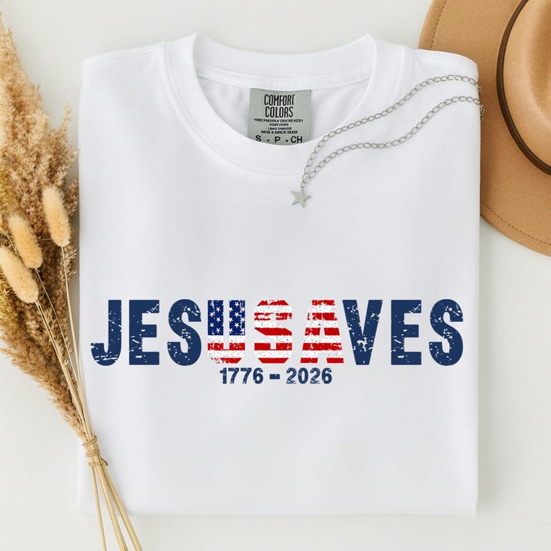 May include: A white t-shirt featuring the text JESUS SAVES in dark blue, with the letters U and S filled with a red, white, and blue American flag pattern. Below the text, the numbers 1776 - 2026 are printed in dark blue. The shirt has a Comfort Colors label.