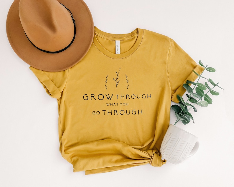 Mustard colored shirt wıth the print on it sayıng "Grow Through What You Go Through" along with flowers on top.