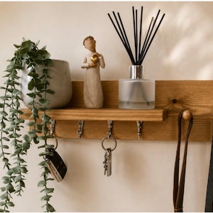 May include: A rectangular light brown wooden wall shelf featuring four silver metal hooks for hanging items. The shelf holds a grey ceramic pot with a trailing green plant, a small beige figurine, and a glass reed diffuser with black sticks.