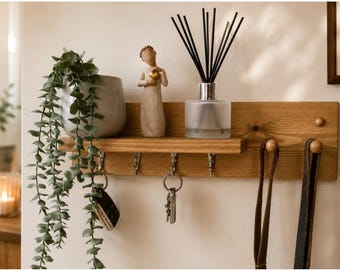 Op de afbeelding: Een rechthoekige wandplank van lichtbruin hout met vier zilverkleurige metalen haken om voorwerpen aan op te hangen. Op de plank staan een grijze keramische pot met een hangende groene plant, een klein beige beeldje en een glazen geurverspreider.