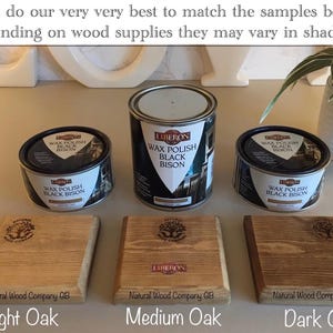 Op de afbeelding: Afbeelding met drie eikenhouten stalen in verschillende tinten, van licht tot donker, met blikken Liberon Wax Polish Black Bison. De stalen zijn gelabeld met Licht Eiken, Medium Eiken en Donker Eiken. De tekst op de afbeelding luidt: "We doen ons uiterste best om de onderstaande voorbeelden te matchen, maar afhankelijk van de houtvoorraad kunnen ze enigszins in tint vari&euml;ren."