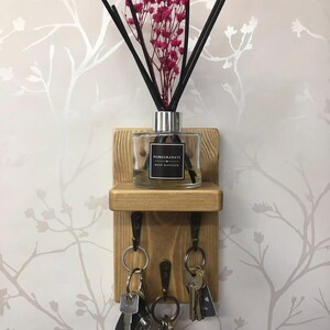 Op de afbeelding: Een houten sleutelhouder met drie haken, een plank en een decoratieve rietverspreider. De diffuser is voorzien van het label "Pomegranate Reed Diffuser" en bevat zwarte rietjes en roze bloemen. Er hangen verschillende sleutels aan de haken.