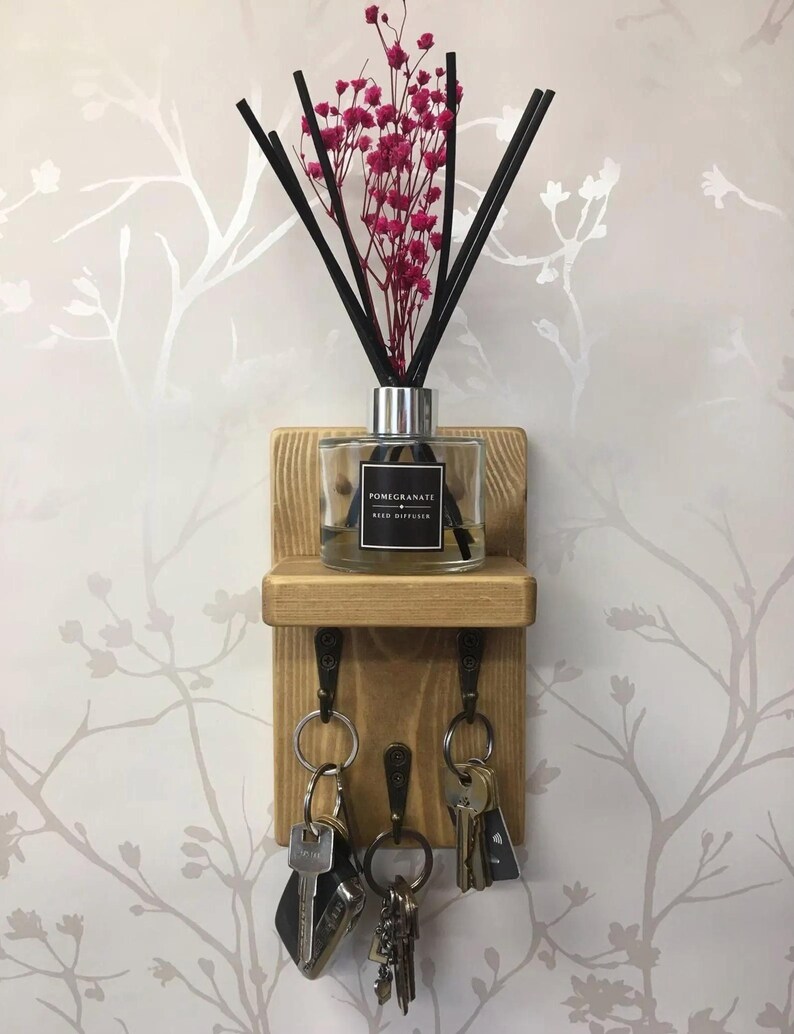 Op de afbeelding: Een houten sleutelhouder met drie haken, een plank en een decoratieve rietverspreider. De diffuser is voorzien van het label "Pomegranate Reed Diffuser" en bevat zwarte rietjes en roze bloemen. Er hangen verschillende sleutels aan de haken.
