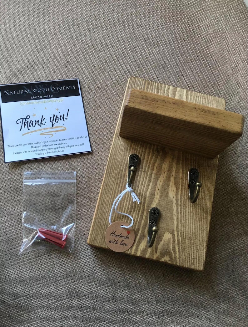 Op de afbeelding: Houten sleutelhouder met drie metalen haken. De sleutelhouder heeft een klein plankje en is gemaakt van lichtgekleurd hout. Een klein label leest "Handmade with love". Een kaart leest "Thank you!" en een klein zakje met rode muurankers is ook inbegrepen.