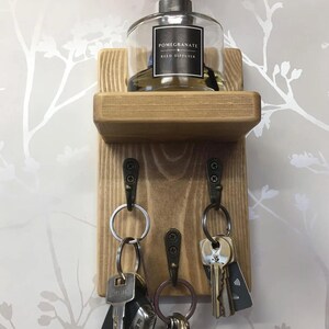 Op de afbeelding: Houten sleutelhouder met een plankje met een glazen fles met het opschrift "Pomegranate Reed Diffuser". Onder het plankje hangen drie metalen haken met sleutels. Het hout heeft een natuurlijke afwerking, tegen een bloemenbehang.