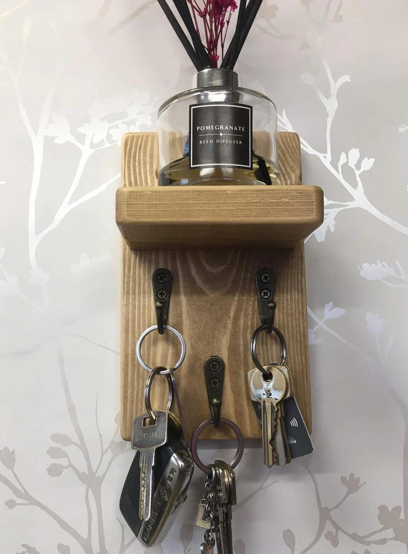 Op de afbeelding: Houten sleutelhouder met een plankje met een glazen fles met het opschrift "Pomegranate Reed Diffuser". Onder het plankje hangen drie metalen haken met sleutels. Het hout heeft een natuurlijke afwerking, tegen een bloemenbehang.