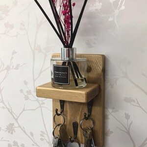 Op de afbeelding: Houten wand sleutelhouder met een klein plankje. Een glazen geurverspreider met zwarte stokjes en roze bloemen staat op de plank. Daaronder drie metalen haken met sleutels. De achtergrond is een bloemenbehang.