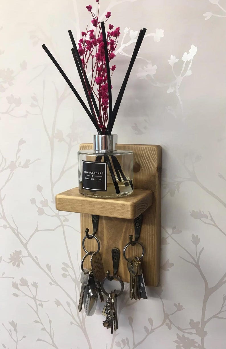 Op de afbeelding: Houten wand sleutelhouder met een klein plankje. Een glazen geurverspreider met zwarte stokjes en roze bloemen staat op de plank. Daaronder drie metalen haken met sleutels. De achtergrond is een bloemenbehang.
