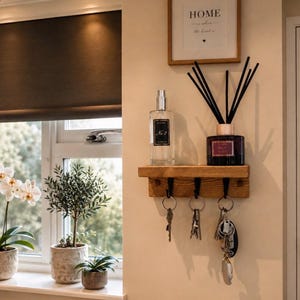 May include: A wall-mounted wooden shelf with three black metal hooks holding sets of keys. On the shelf sits a clear glass bottle labelled No. 3 and a dark glass reed diffuser bottle labelled No. 7 with black reeds. Above hangs a framed sign reading HOME.