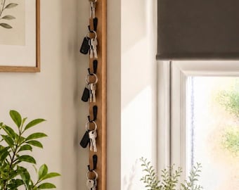 Op de afbeelding: Een verticaal houten wandrek voor sleutels met zes gelijkmatig verdeelde zwarte metalen haken. Elke haak houdt een metalen sleutelhanger met diverse sleutels vast. De lichtgekleurde houten strip is vlak tegen een binnenmuur bevestigd.