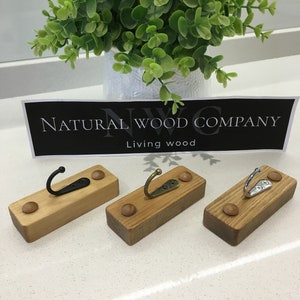 May include: Three wooden wall hooks with metal hooks and decorative wooden pegs. The hooks have different metal finishes: black, bronze, and silver. The hooks are rectangular and made of light-coloured wood. The sign says "NATURAL WOOD COMPANY Living wood".