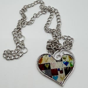 May include: Silver-toned necklace with a heart-shaped pendant. The pendant features a collage of colorful heart designs. The chain is a classic link style, and the pendant has a decorative silver border.