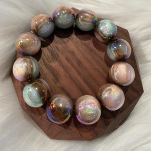 May include: A circular bracelet composed of twelve spherical beads. Each bead features a smooth, glossy finish with a swirling iridescent effect, blending shades of metallic copper, soft pink, and pale blue with a reflective, pearlescent sheen.