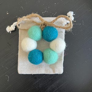 May include: A small wreath made of felt balls in white, teal, and light green colors. The wreath is in a small, white drawstring bag with a brown twine bow.
