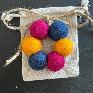 May include: A small pouch with a drawstring closure containing a set of six felt balls in a circular arrangement. The balls are in three colors: yellow, pink, and dark blue.