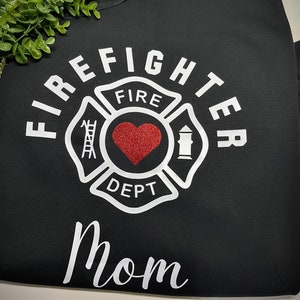 May include: Black t-shirt with a white firefighter emblem and the word "FIRE DEPT" inside. A red heart with glitter is in the centre of the emblem. The word "FIREFIGHTER" is written around the emblem and the word "MOM" is written below the emblem.