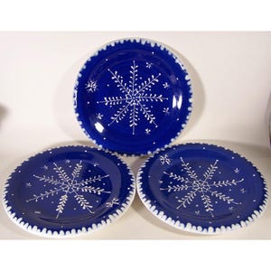 May include: Three blue ceramic plates with white snowflake designs. Each plate has a white scalloped edge. The snowflakes are hand-painted with delicate details, creating a festive winter theme.